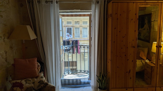A picture from inside a dark room of the balcony across the street, with an old woman hanging out the window smoking above a line of clothes drying