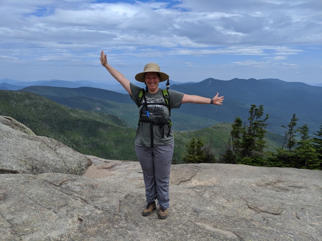 A woman wearing a straw hat at the top of a mountain with her arms in a weird victory pose.