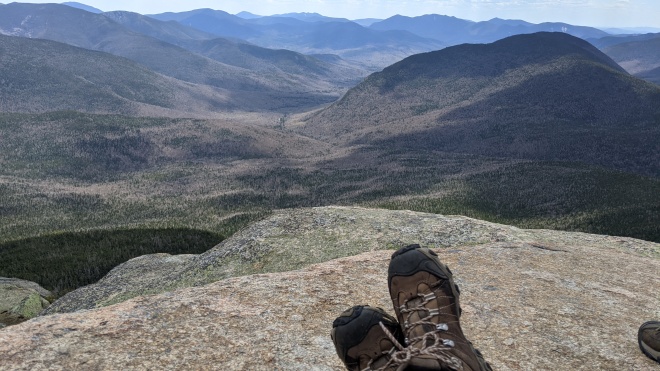 A pair of hiking boots on a granite boulder overlooking a mountain valley with a range of mountains around it and a solo mountain in the middle