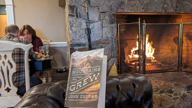 The book "How the Mountains of North America Grew" centrally, with a fire to the right, two people playing GO to the left, and glimpses of a leather couch